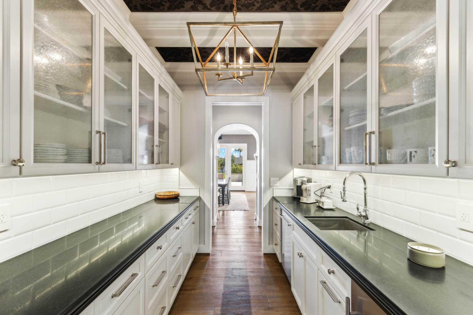 A butler’s pantry lined on both sides with white cabinetry, glass-front upper cabinets, dark stone counters, a secondary sink, and additional prep and storage space.