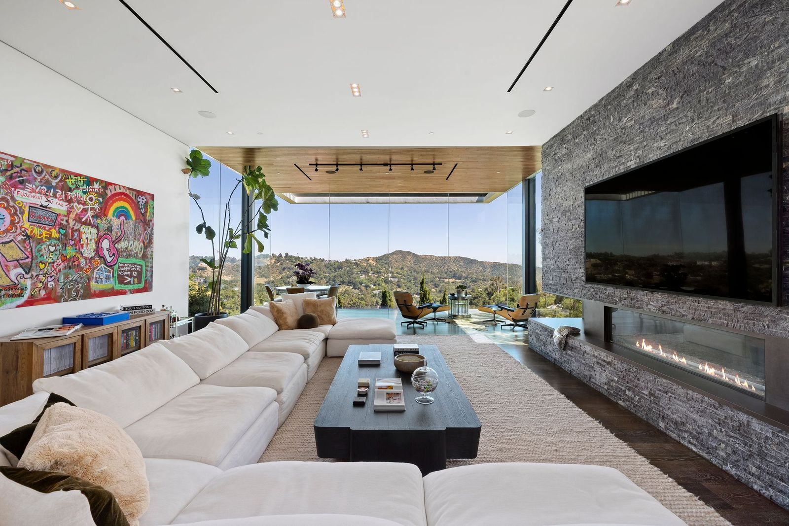 Before reaching the glass walls, a large sectional fills the main seating area opposite a stacked-stone fireplace wall, where a linear fireplace and mounted television are built into the dark textured surface.
