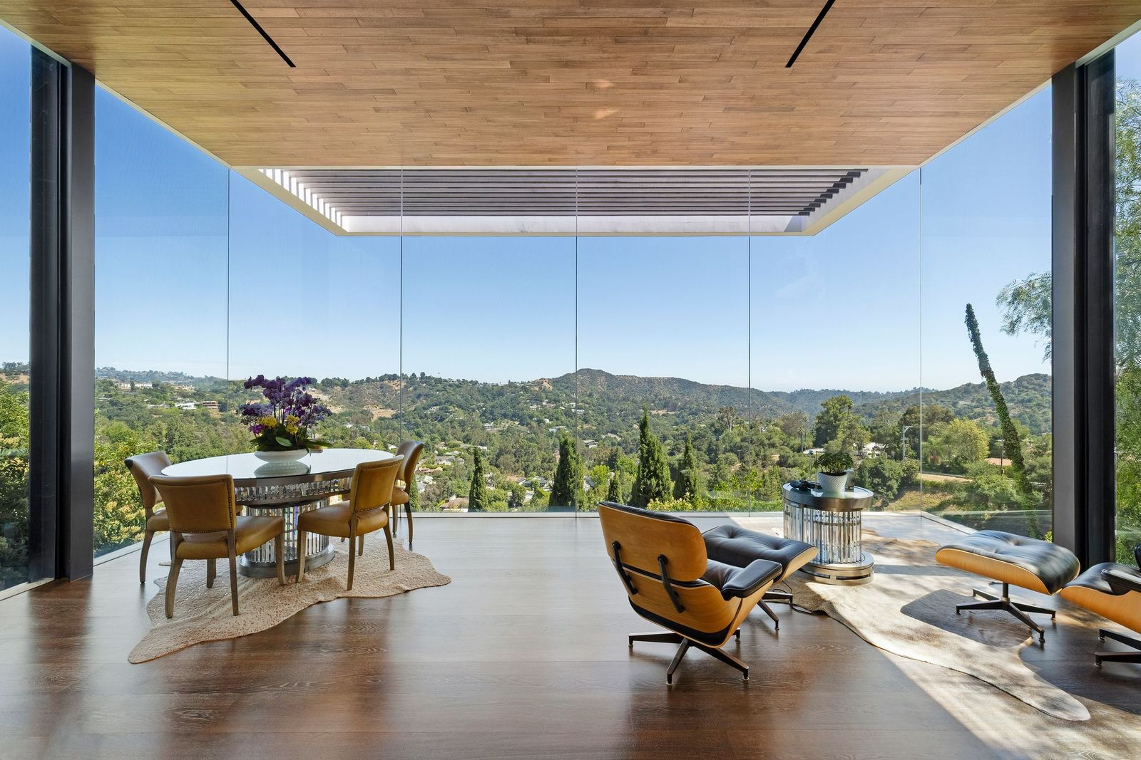 The room narrows into a second seating zone beneath a wood-paneled ceiling, furnished with lounge chairs and a round table positioned directly against the glass. 