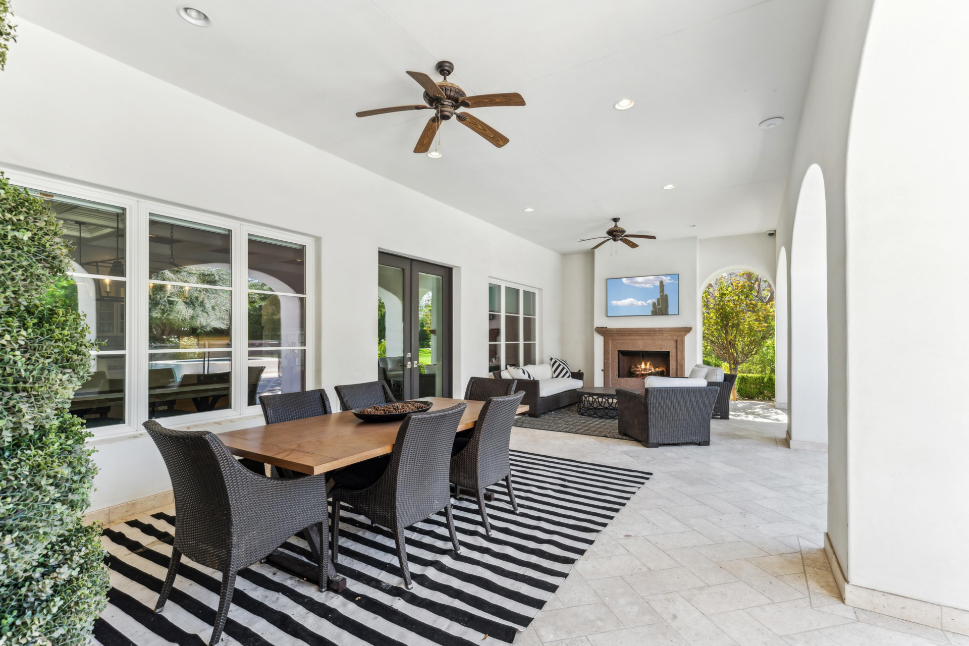 Stone flooring travels across the full length of the space, with one side set up for dining and the other arranged around an outdoor fireplace and television.