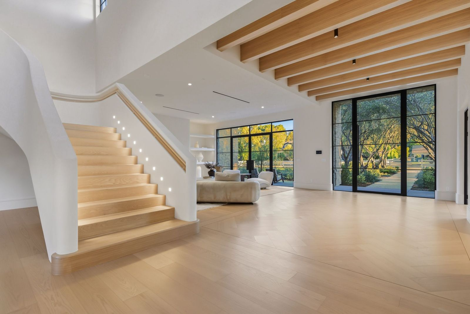 Beside the sitting area, a floating white oak staircase rises through the main living spaces, its sculpted plaster walls curving around the steps while small lights trace the steps.