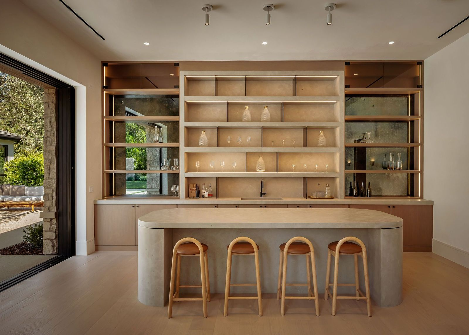 An oversized stone-topped island runs through the center with seating for four, while the back wall combines illuminated open shelving, mirrored panels, cabinetry, a sink, and built-in bottle storage.