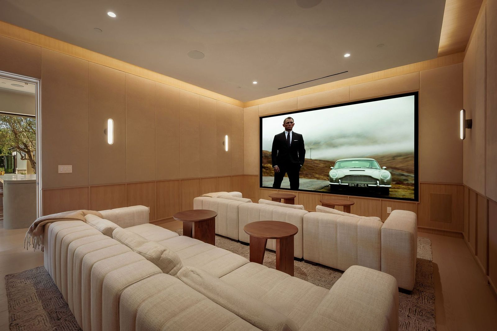 The professional theater is wrapped in upholstered wall panels and wood wainscoting with soft perimeter ceiling lighting, while two rows of deep white couches face a wall-filling projection screen.