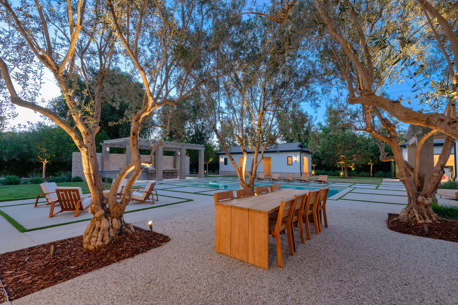 A separate patio opens beneath a grove of olive trees where a long wood dining table creates an outdoor dining area.