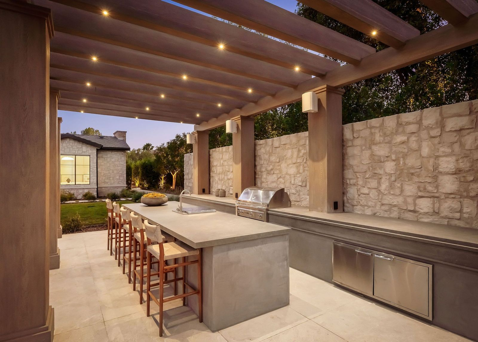 A long counter provides bar seating for several guests, with an integrated grill, sink, refrigeration, and additional stainless-steel storage behind it.
