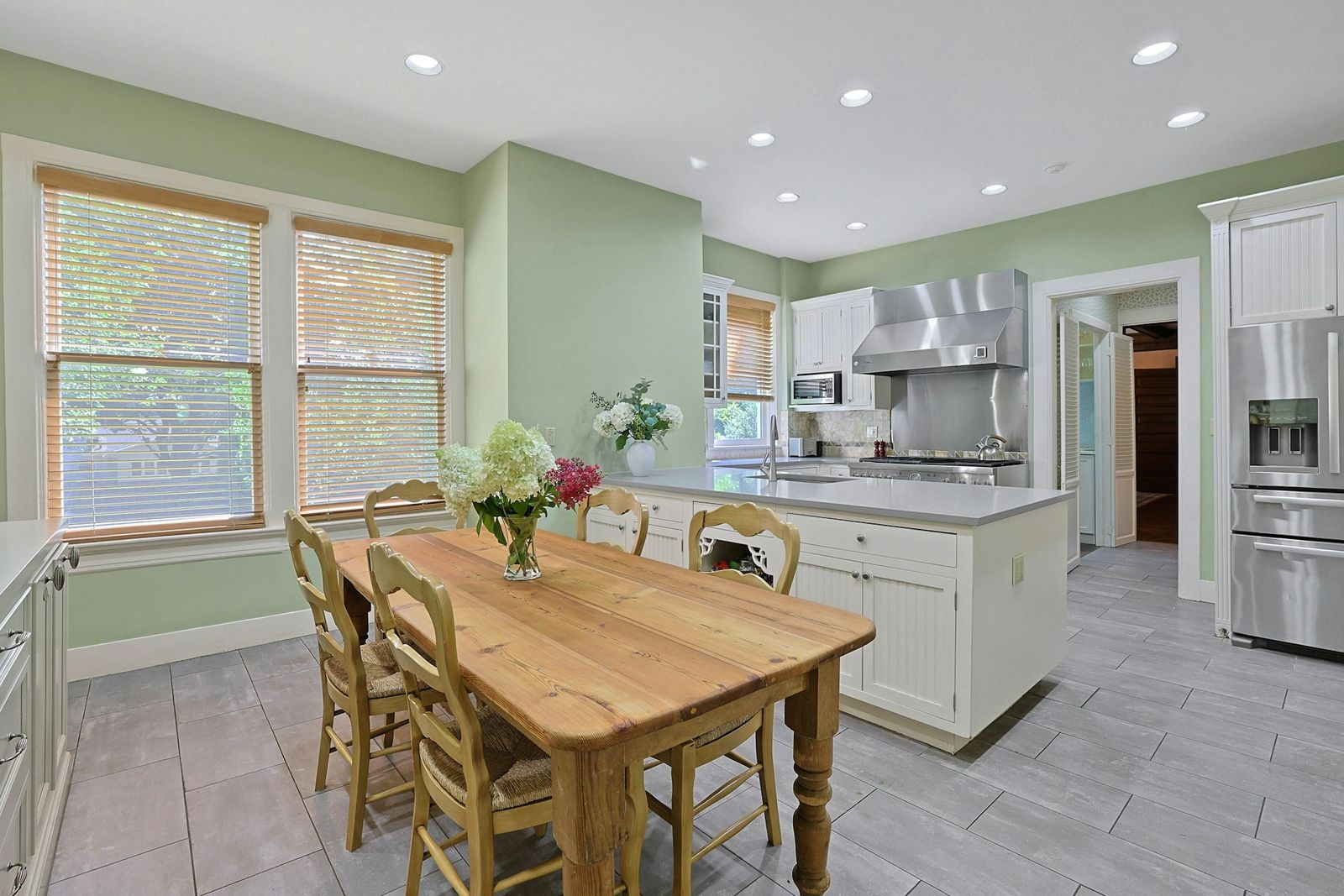 The kitchen begins with a casual dining area set beside a pair of windows, made up of a large wooden table and five chairs. 