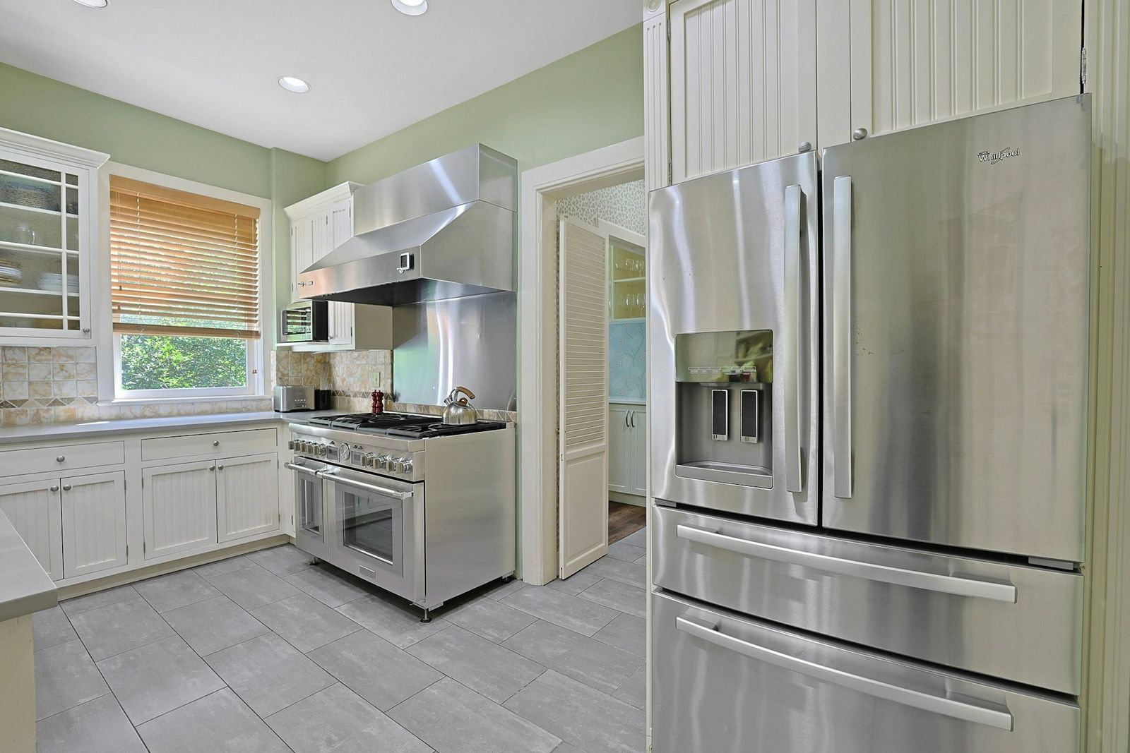 White cabinetry lines the walls around long counters, while a professional-style stainless-steel range sits beneath an oversized hood and a full-size refrigerator is built into the opposite wall.