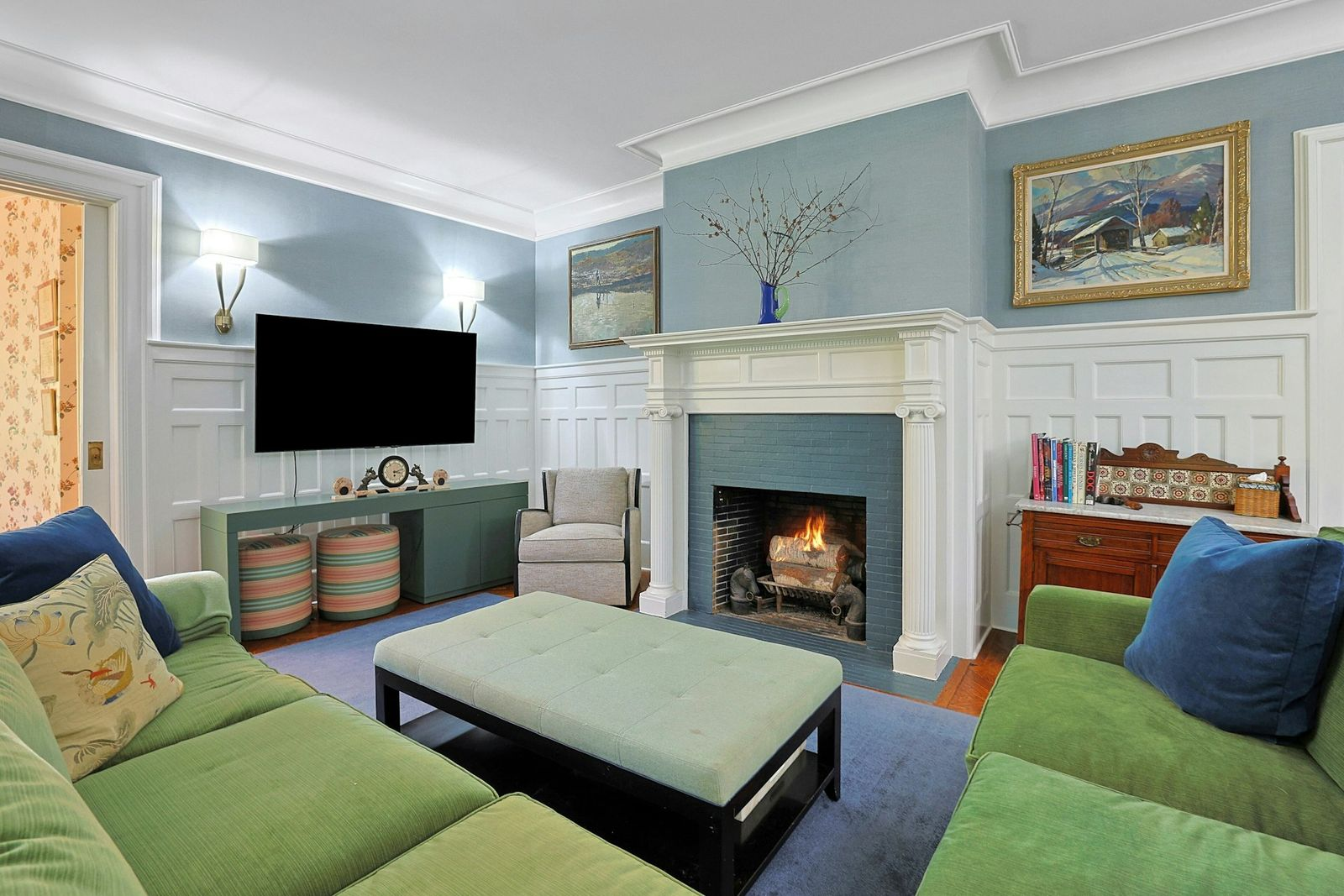 The family room where a pair of green sofas are arranged around a large upholstered ottoman facing the fireplace. 