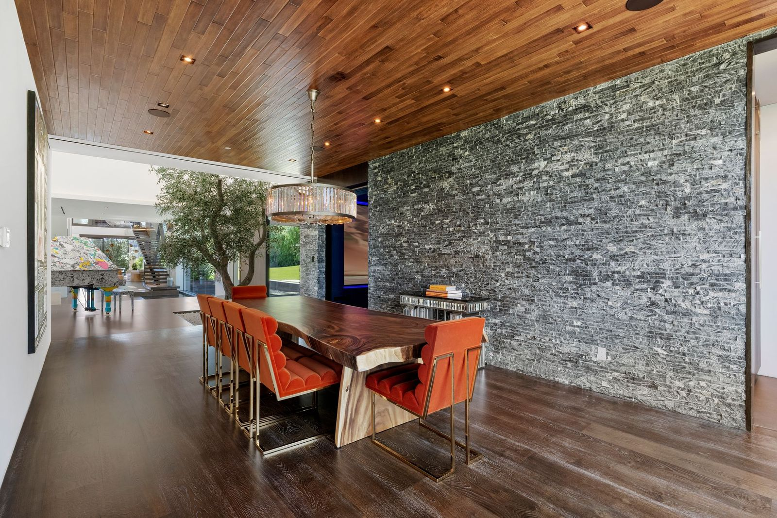 A live-edge dining table runs through the center beneath a circular crystal chandelier, with dark orange velvet chairs adding contrast against the gray stone and dark timber.