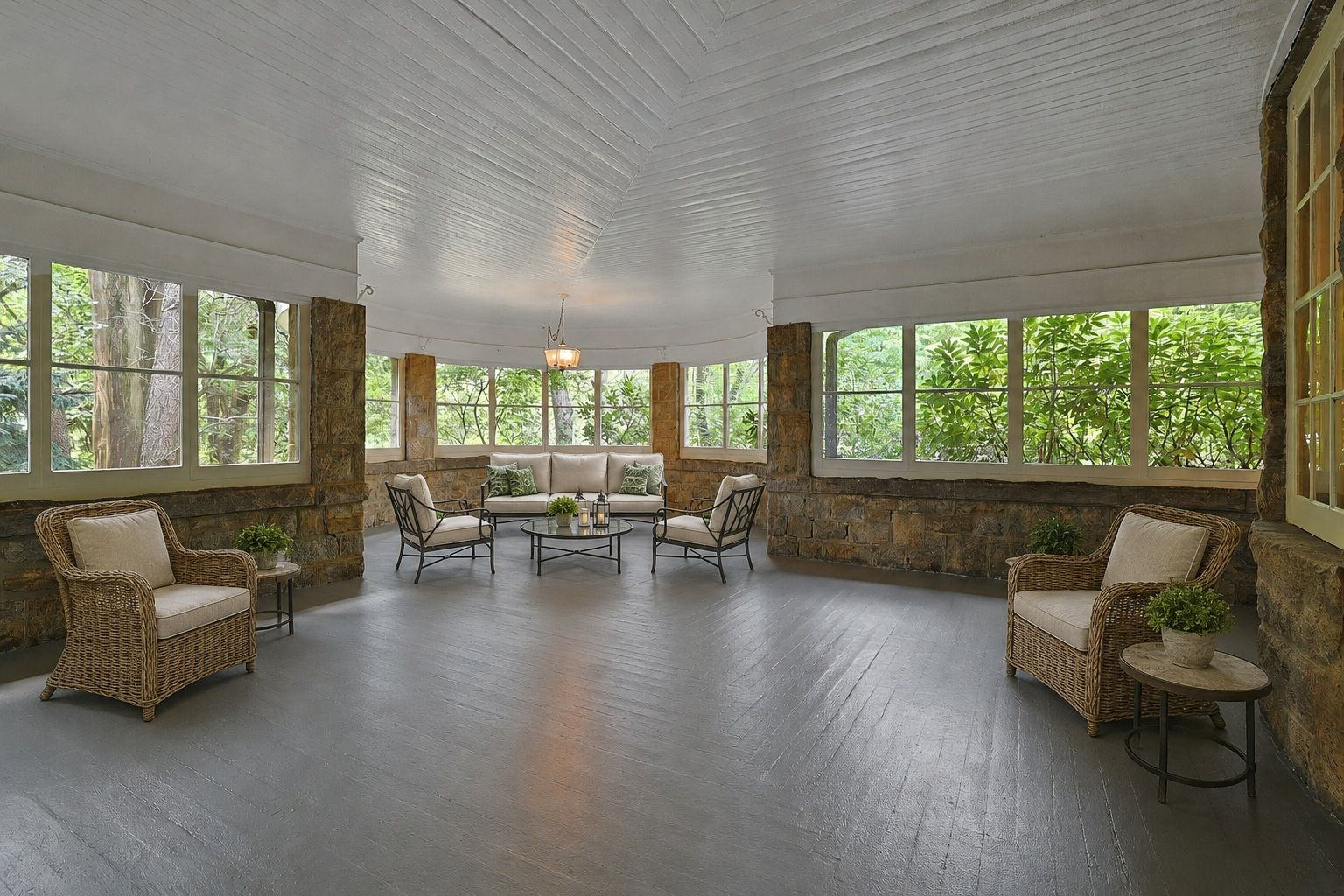 Stone walls and columns frame long runs of windows around the perimeter, while several seating arrangements are spread across the porch, including wicker chairs along the sides and a larger lounge within the curved part of the porch.
