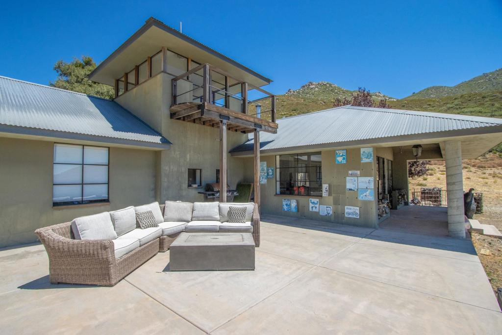 A large concrete patio creates an open gathering area with lounge seating before the entrance moves beneath a covered porch supported by thick cylindrical columns.