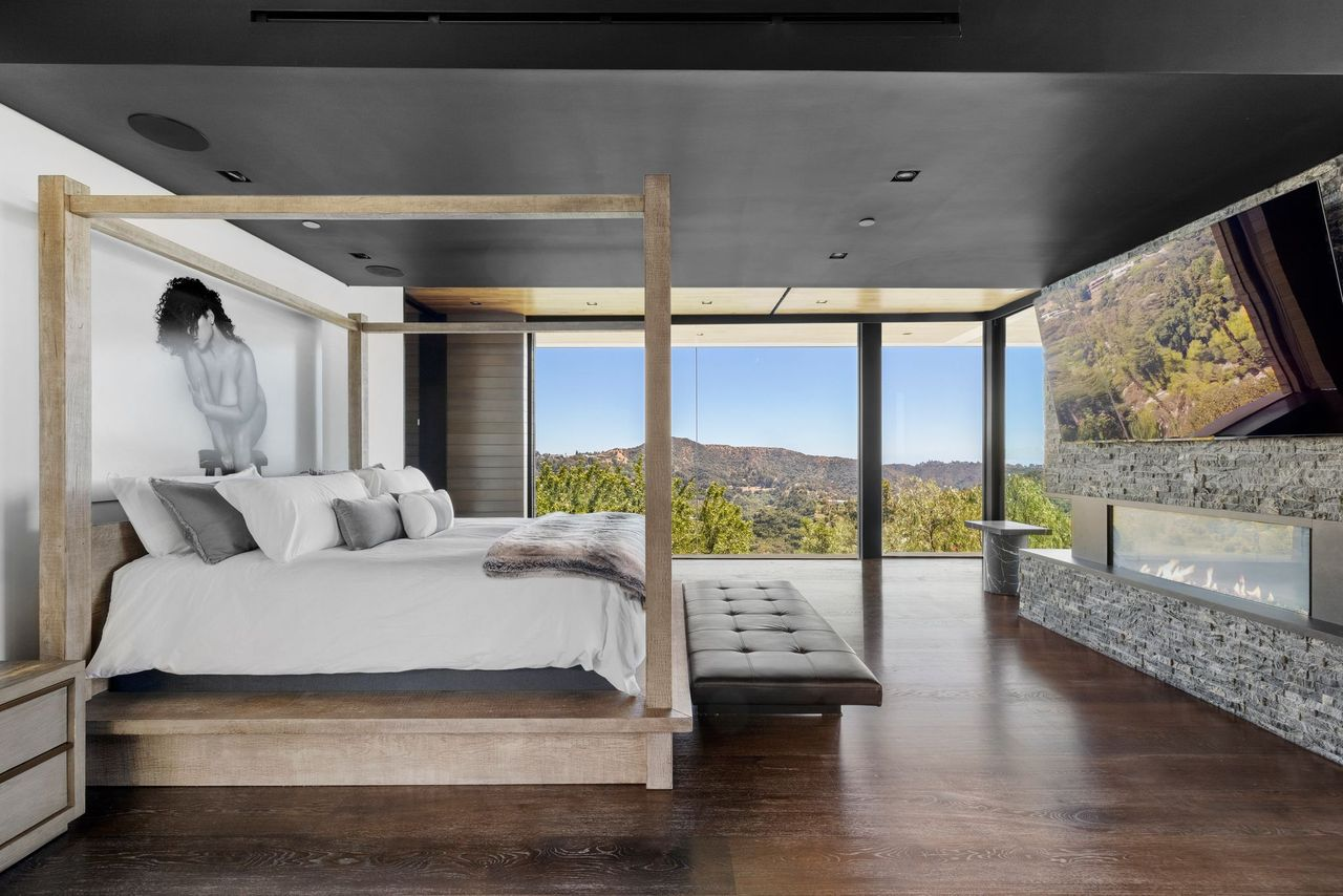 A pale wood four-poster bed sits against one side, while a long stacked-stone fireplace runs opposite it beneath a mounted television.