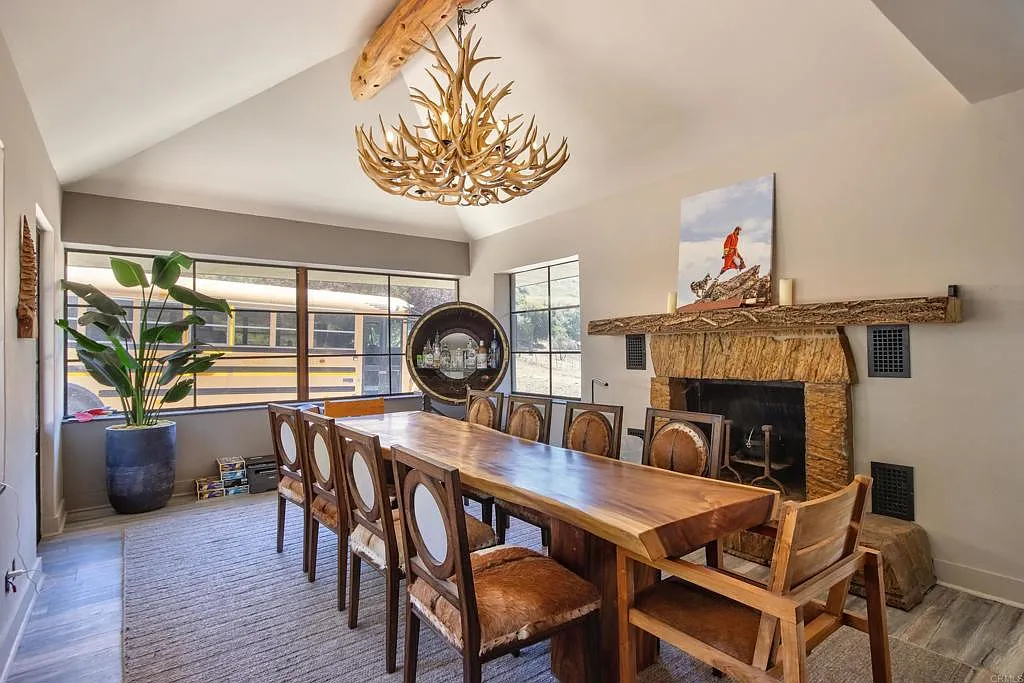 The dining room has a long live-edge wood table with seating for a large group beneath an antler-style chandelier, and a stone fireplace next to it.