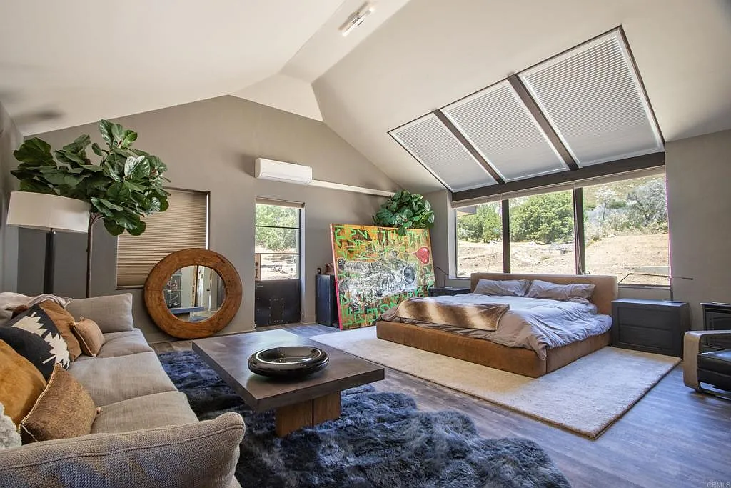 The primary bedroom sits beneath another vaulted ceiling, with the bed positioned against a long wall of windows and across from a lounge area with a plush couch.