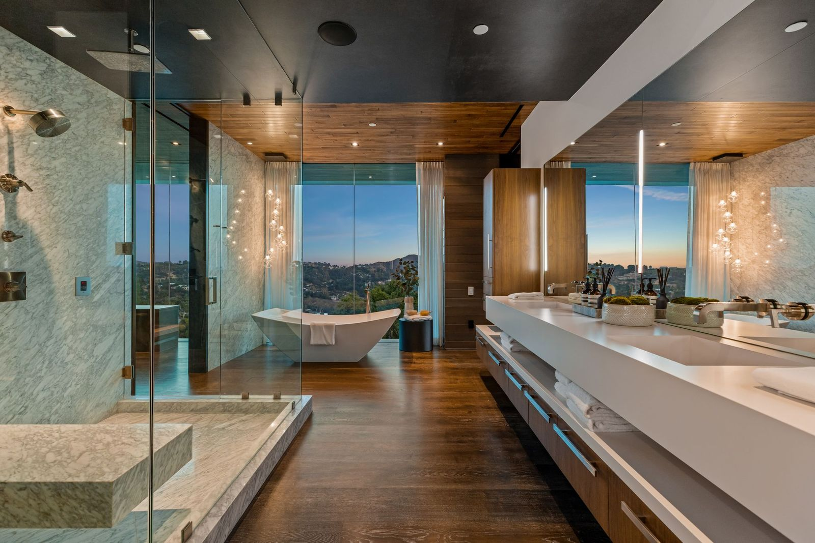 A long floating vanity runs along one wall beneath an expansive mirror, while the opposite side is occupied by a glass-enclosed shower with a built-in stone bench and wall-mounted fixtures.