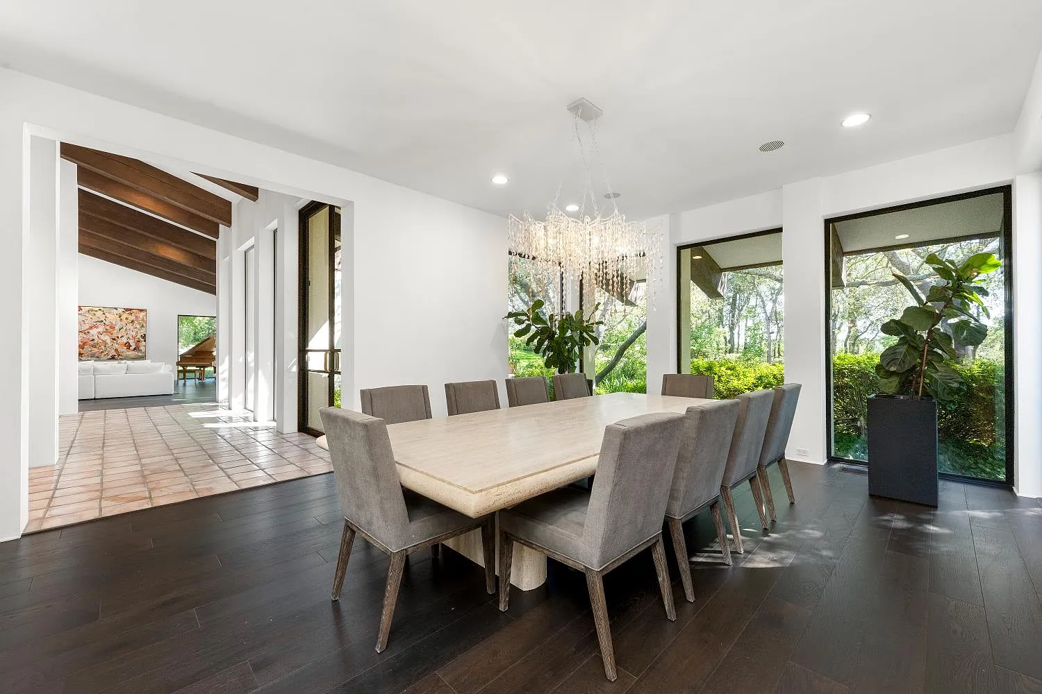 The formal dining room features a long rectangular table beneath a beaded chandelier, with floor-to-ceiling windows looking out to the greenery surrounding the home.
