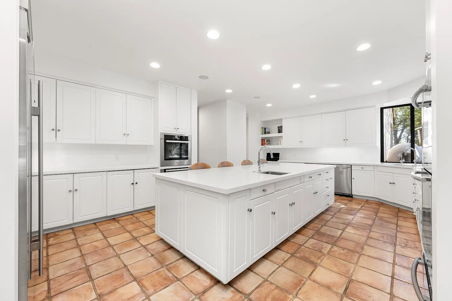 The kitchen is arranged around a large central island, with white cabinetry wrapping the perimeter and stainless-steel appliances built into the surrounding walls.