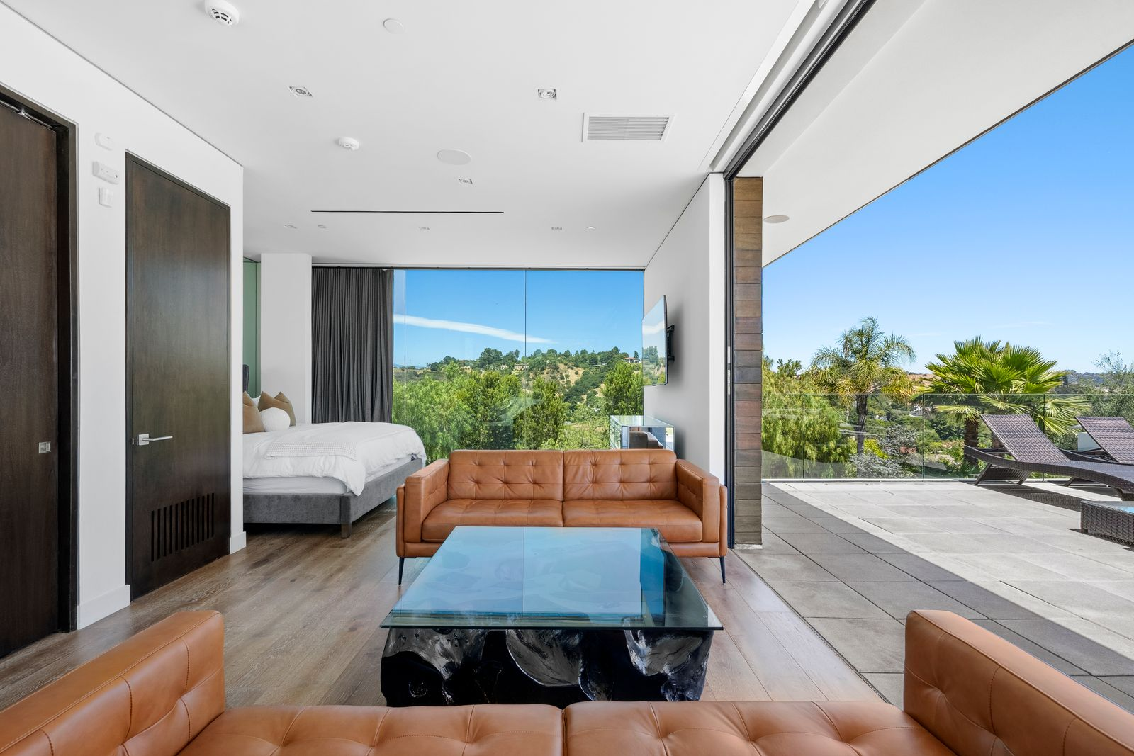 Inside, the bedroom is arranged beside a compact sitting space furnished with caramel leather sofas, while floor-to-ceiling glass opens the room directly to the surrounding hillside. 