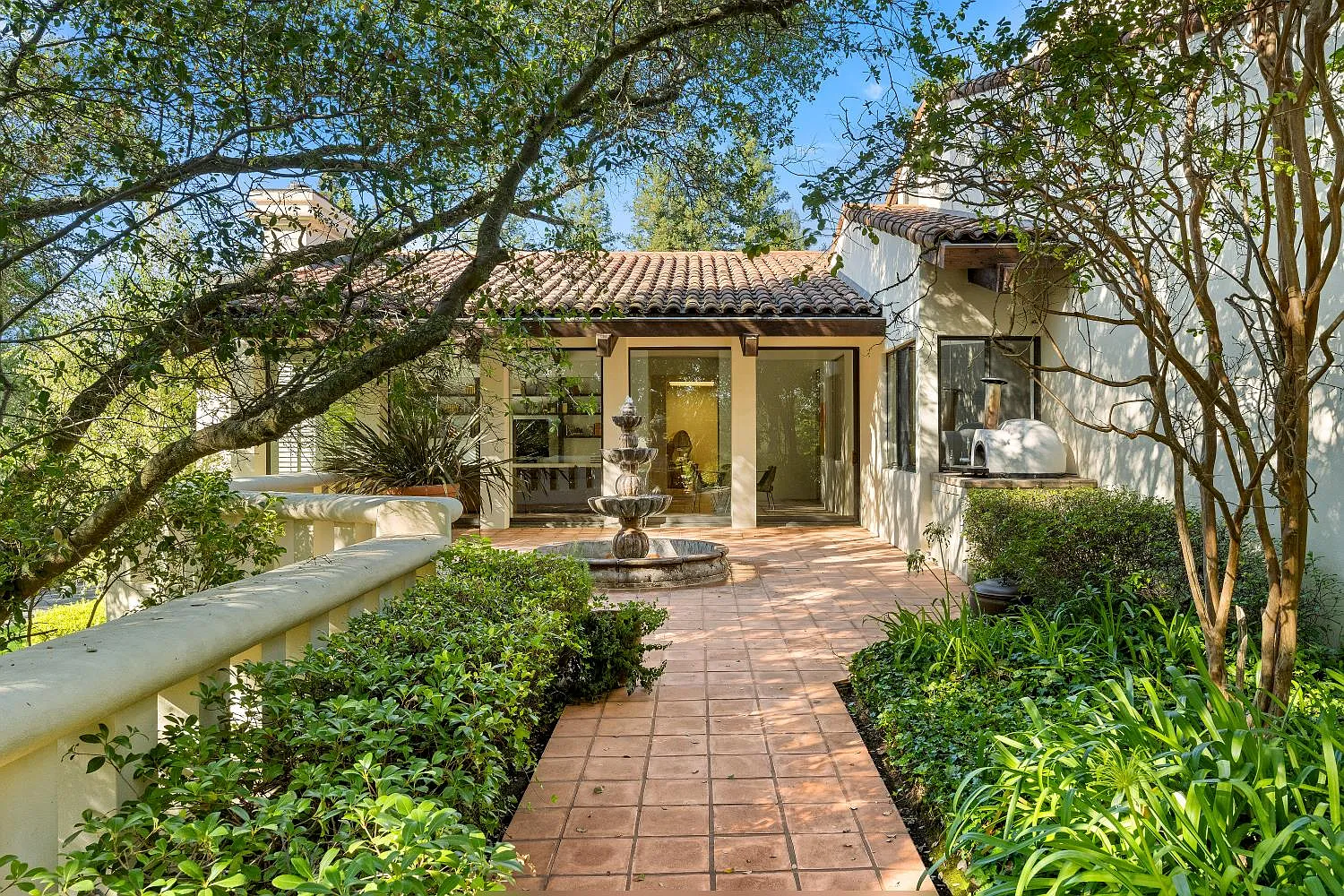 A terracotta-tiled courtyard surrounded by greenery has a tiered fountain in the center and a built-in pizza oven tucked along the wall beside the house.