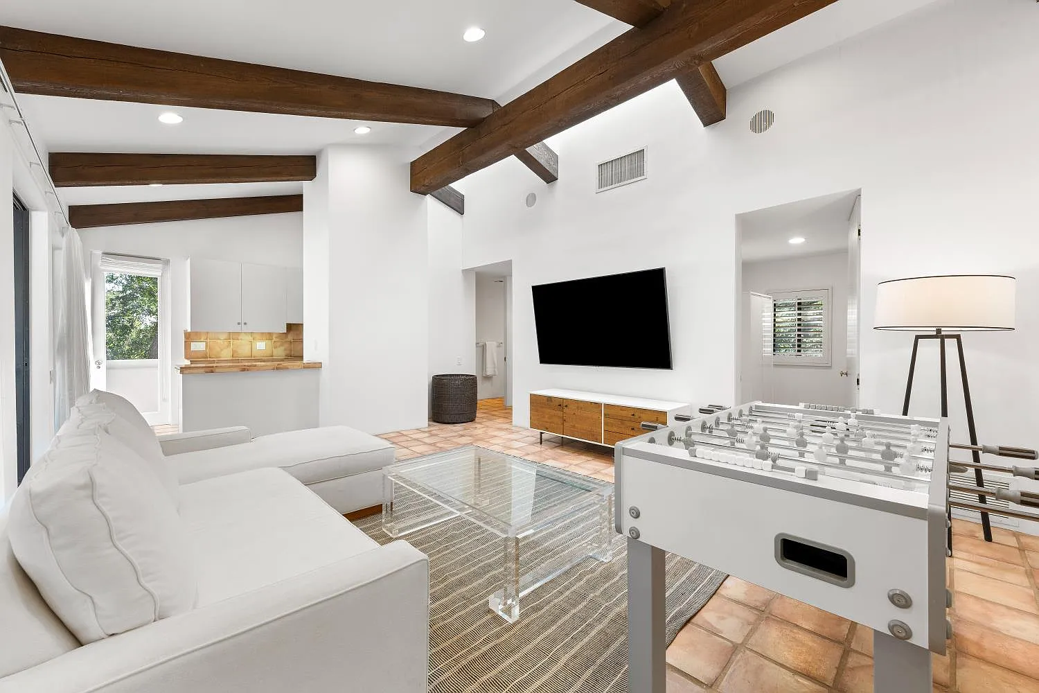 Living room is arranged beneath exposed wood beams with a sectional facing the television and foosball table, while a compact full kitchen sits on the other side of the room.