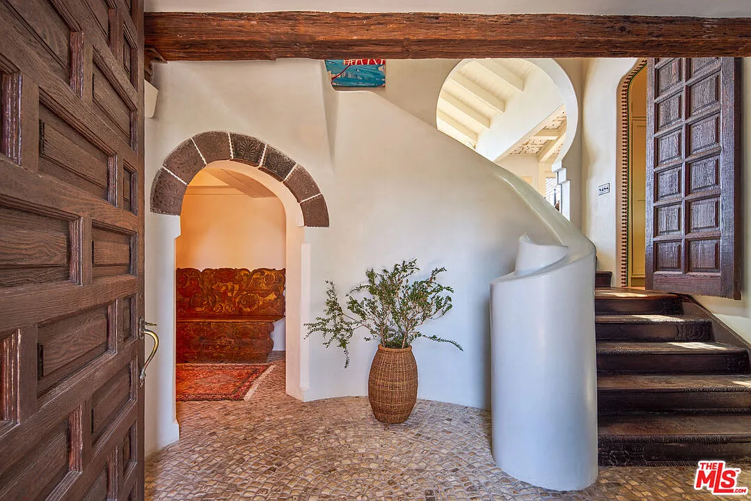 A heavy paneled wood door opens to a curved plaster staircase that sweeps upward along the wall, while a smaller arched opening framed in dark stone leads into another part of the house.