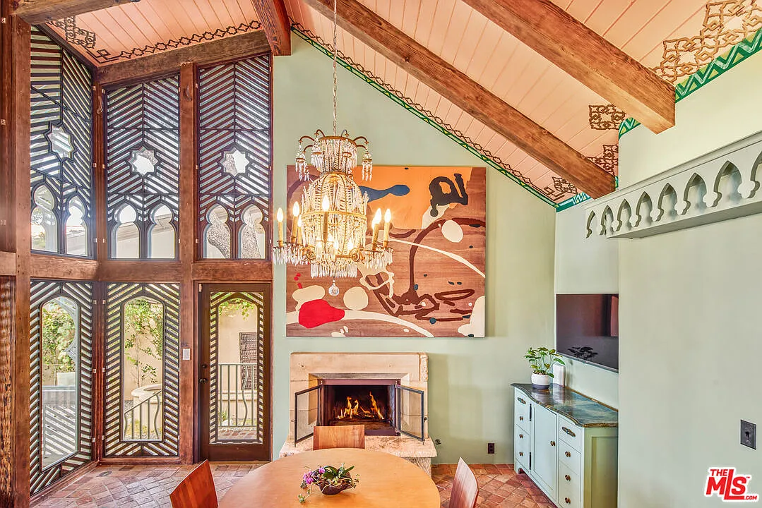 A round table sits beside a fireplace, with tall patterned windows and dark wood framing bringing in light across from a mounted TV.