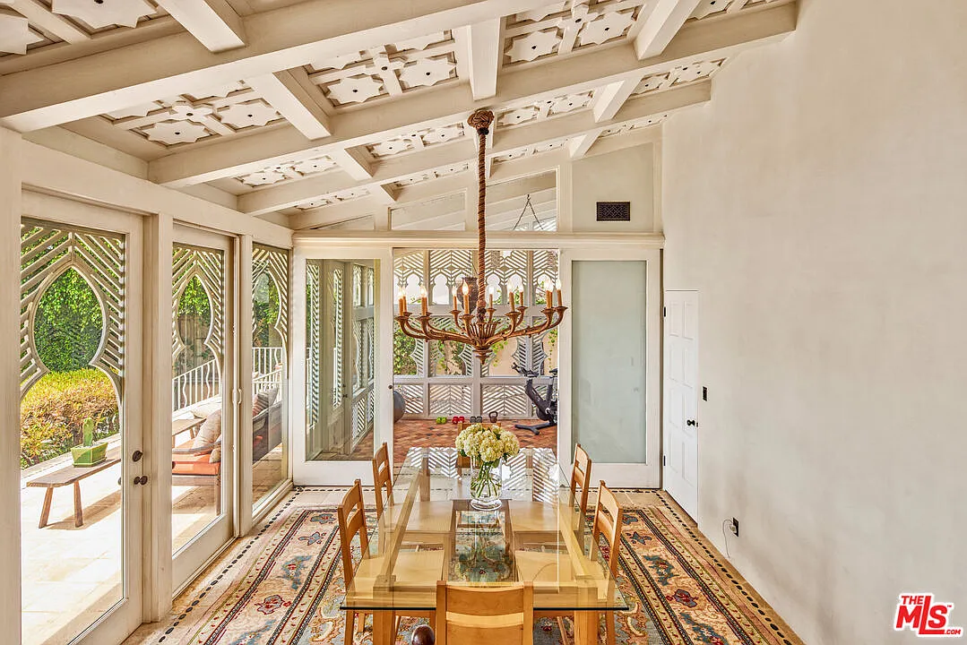 A long glass-topped table runs down the center beneath a sculptural chandelier, while tall patterned windows and glass doors line one side of the room.