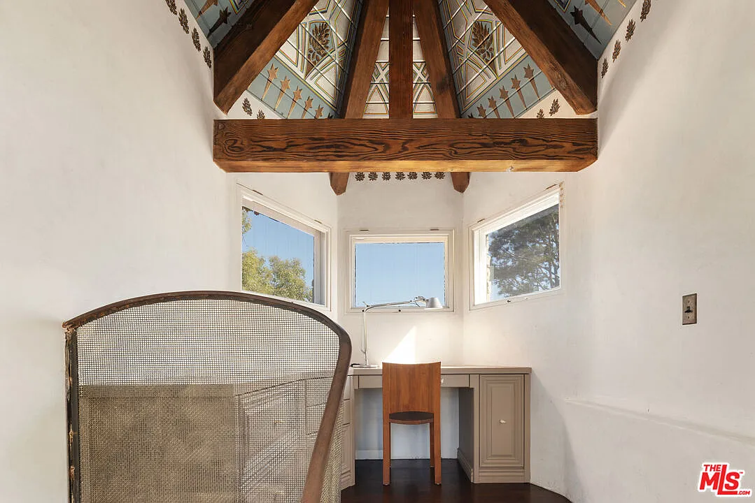 The compact room sits beneath a steeply pitched, exposed-beam ceiling, with painted geometric detailing above three ocean facing windows and a built-in desk.