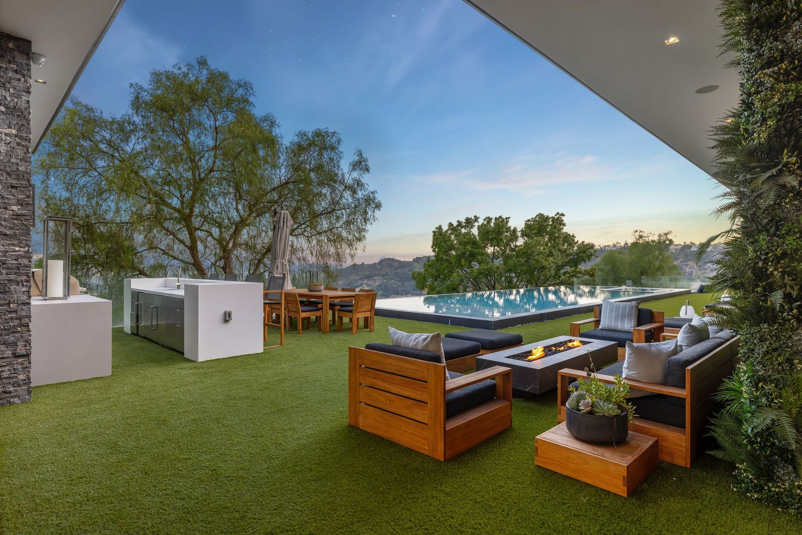 Beneath the home’s overhang, a seating area gathers around a rectangular fire table, while a nearby outdoor kitchen adds stainless-steel cabinetry, a grill, and a separate dining table positioned closer to the pool.