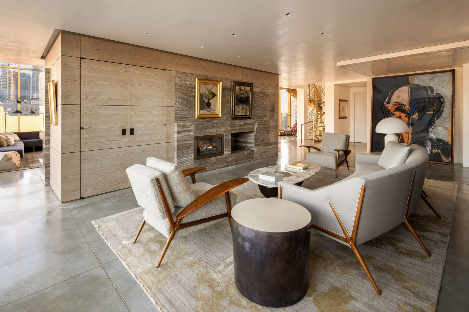 Facting the fire, low-profile sofas and sculptural wood-framed chairs gather around a stone-topped coffee table, while open passages on either side keep the room sunlit from the walls of glass.