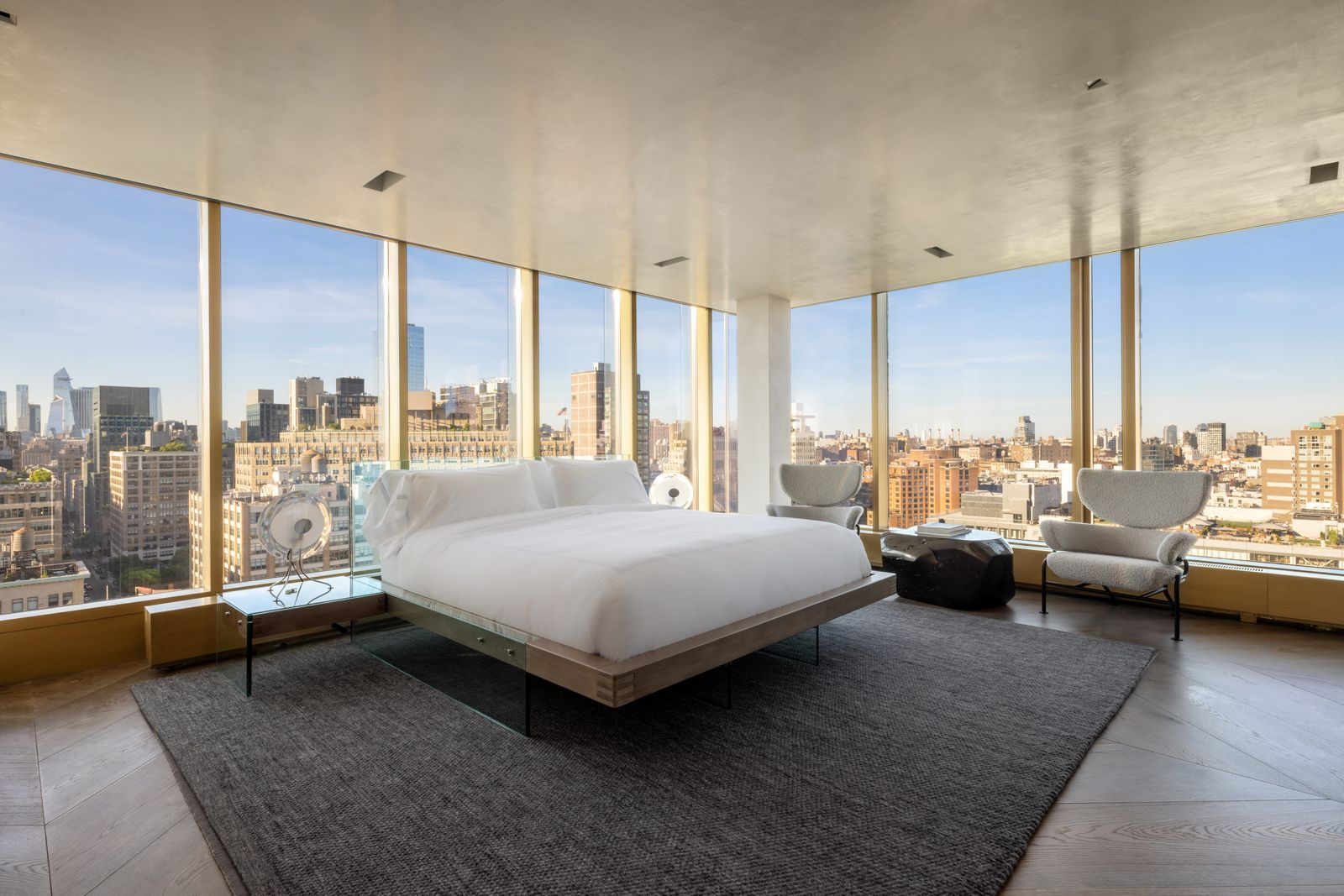 Floor-to-ceiling windows wrap around two sides of the bedroom, placing the bed directly against the city backdrop. 