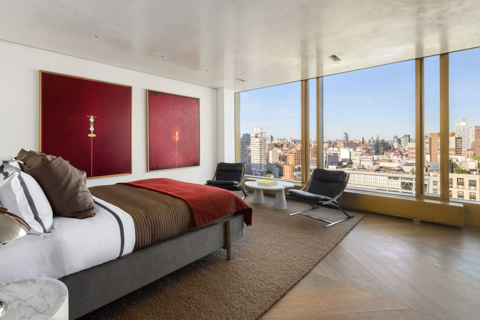 Two deep red artworks fill the wall between the single bed and a pair of low black lounge chairs sitting beside the floor-to-ceiling windows.