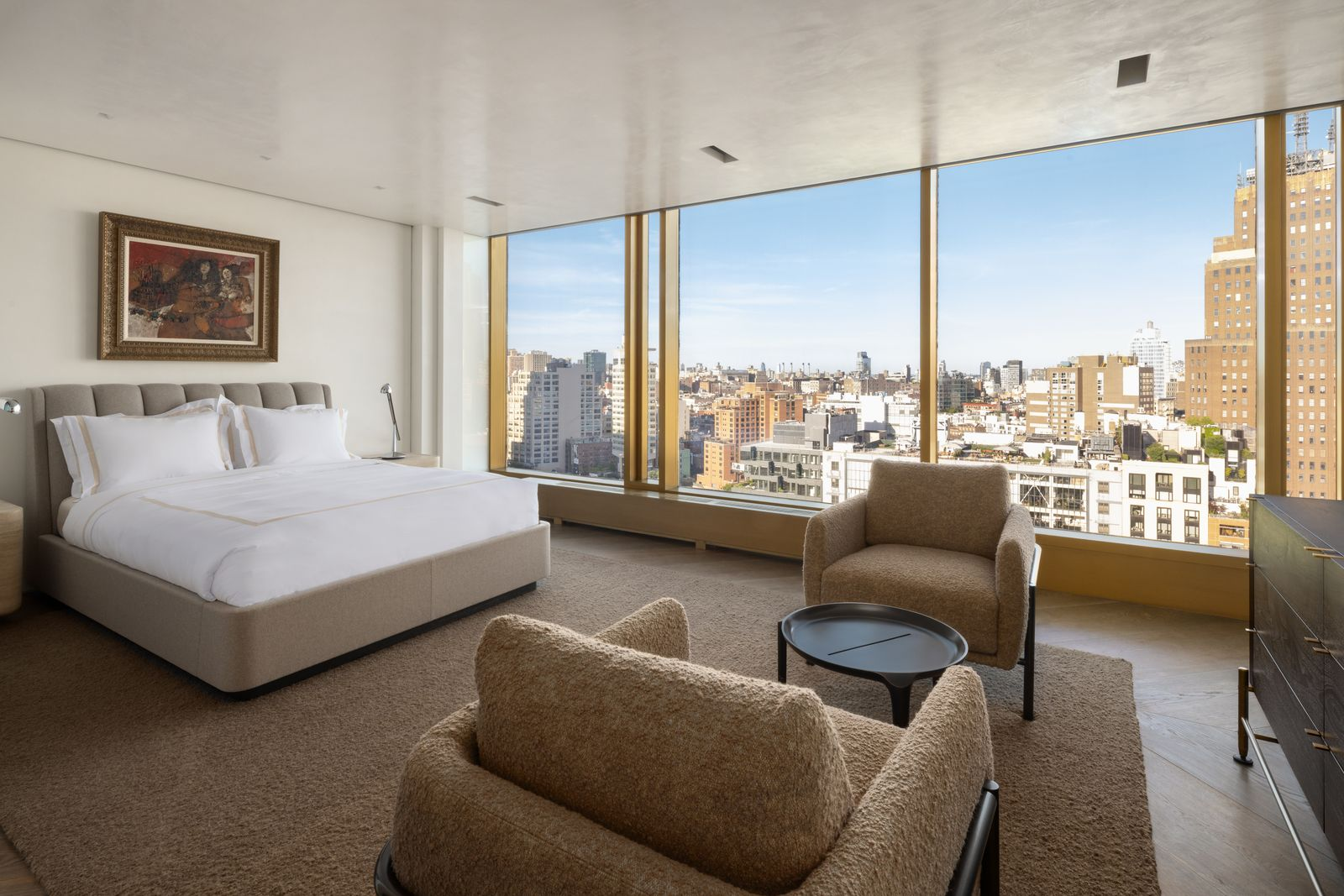 The second bedroom has a much softer color palette against the skyline, with an upholstered bed and cushioned armchairs all arranged down the center of the room.