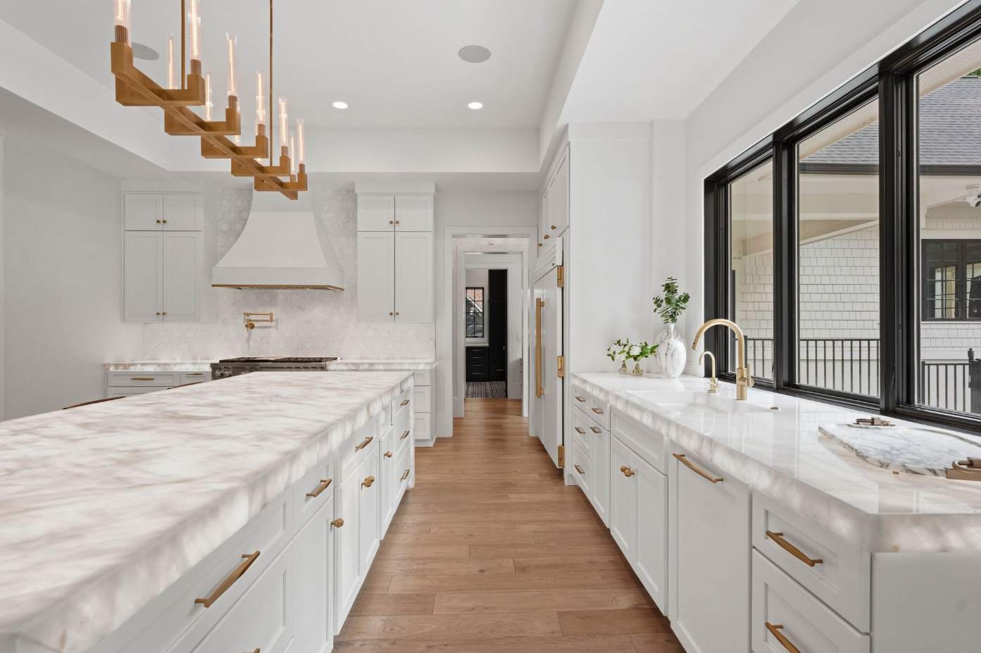 White custom cabinetry extends across the surrounding walls with brass hardware, framing a full suite of Wolf appliances, concealed refrigeration, and a broad range beneath a custom hood. 