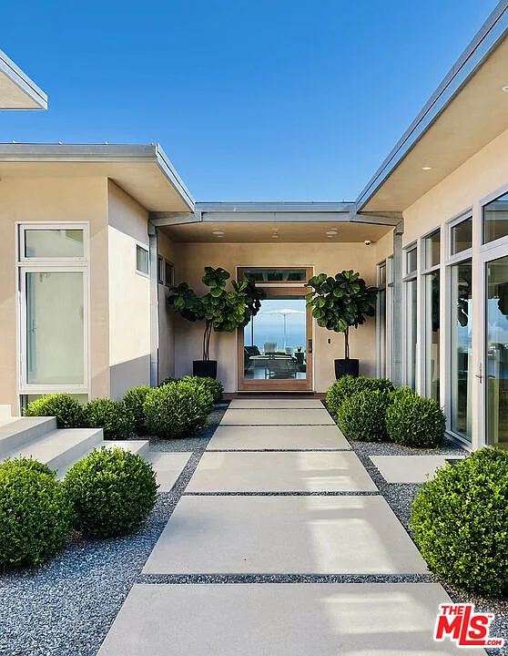 Rounded boxwood plants line both sides, softening the precision as the path moves between the home’s low, flat-roofed wings to the glass front door with a view of the ocean.