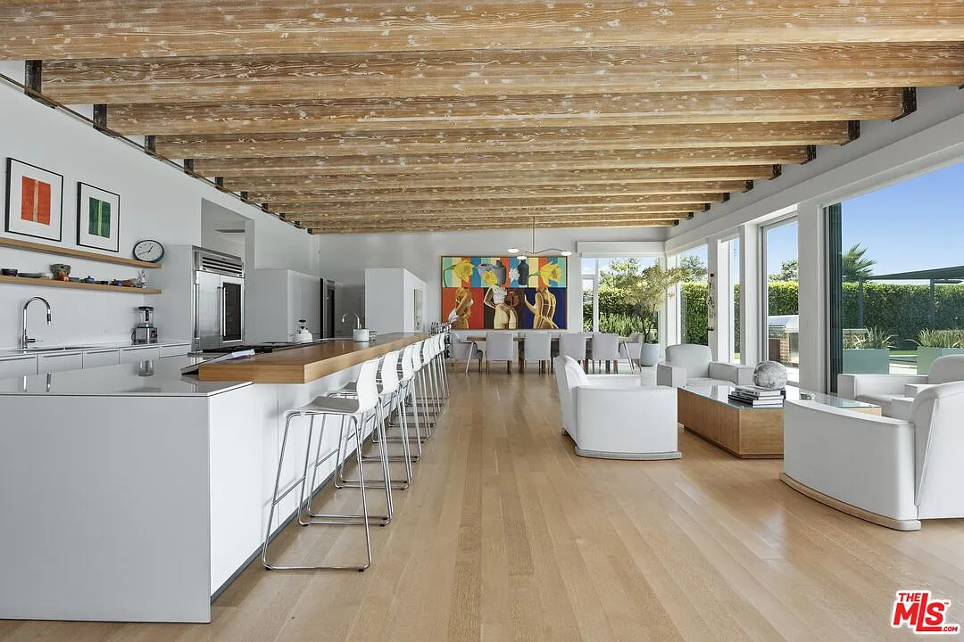 Beyond the kitchen, the dining area places a long table beneath the same exposed-beam ceiling, backed by a large colorful artwork and glass doors opening toward the landscaped side terrace.
