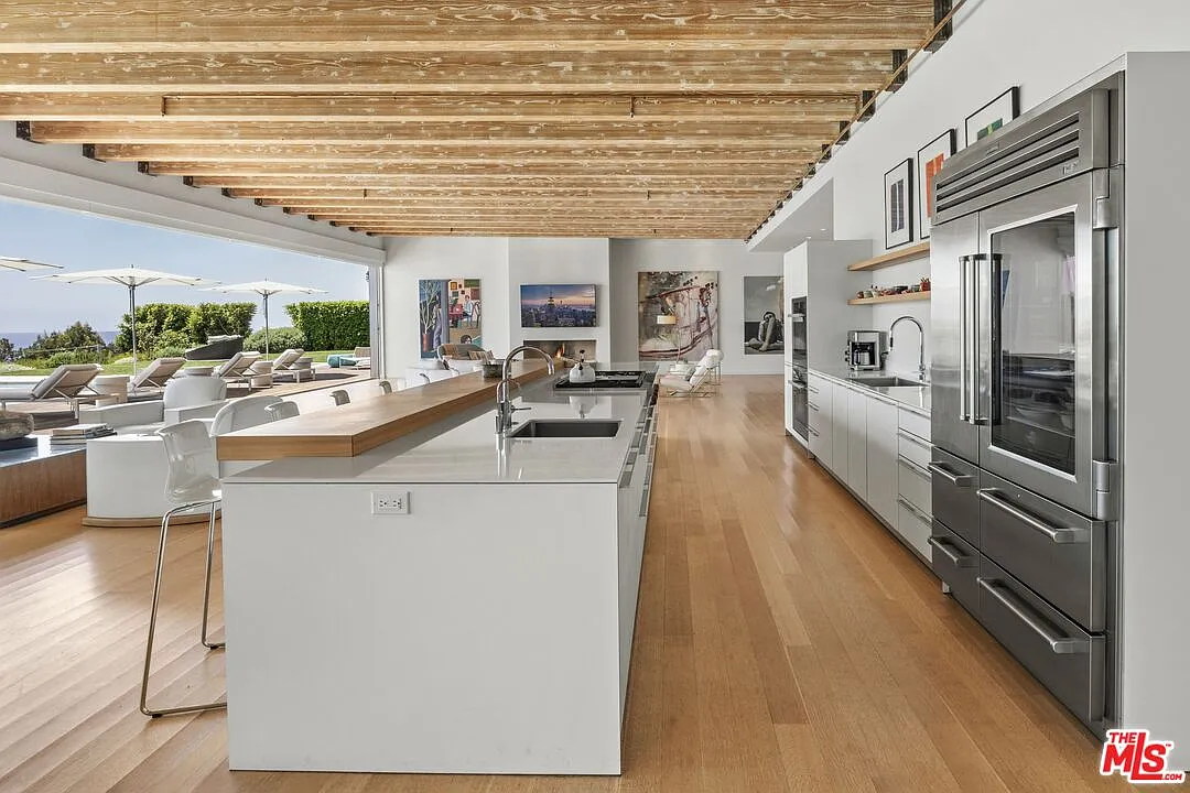 A long white island features one of two sinks and the stovetop, while a raised wood counter runs along its side to create an extended breakfast bar with bartop seating.