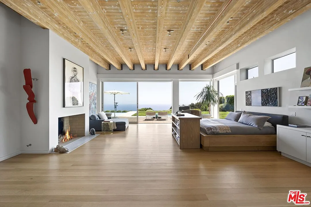 The bedroom stretches beneath weathered wood beams, with the bed positioned right across from the fireplace.