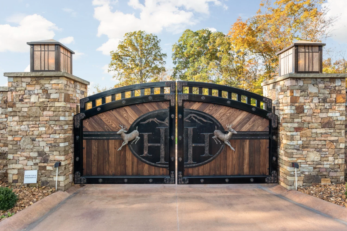 Entering Hidden Hollow Ranch begins at a pair of wood-and-iron gates set between tall stone pillars, opening to a long private drive through the wooded acreage.