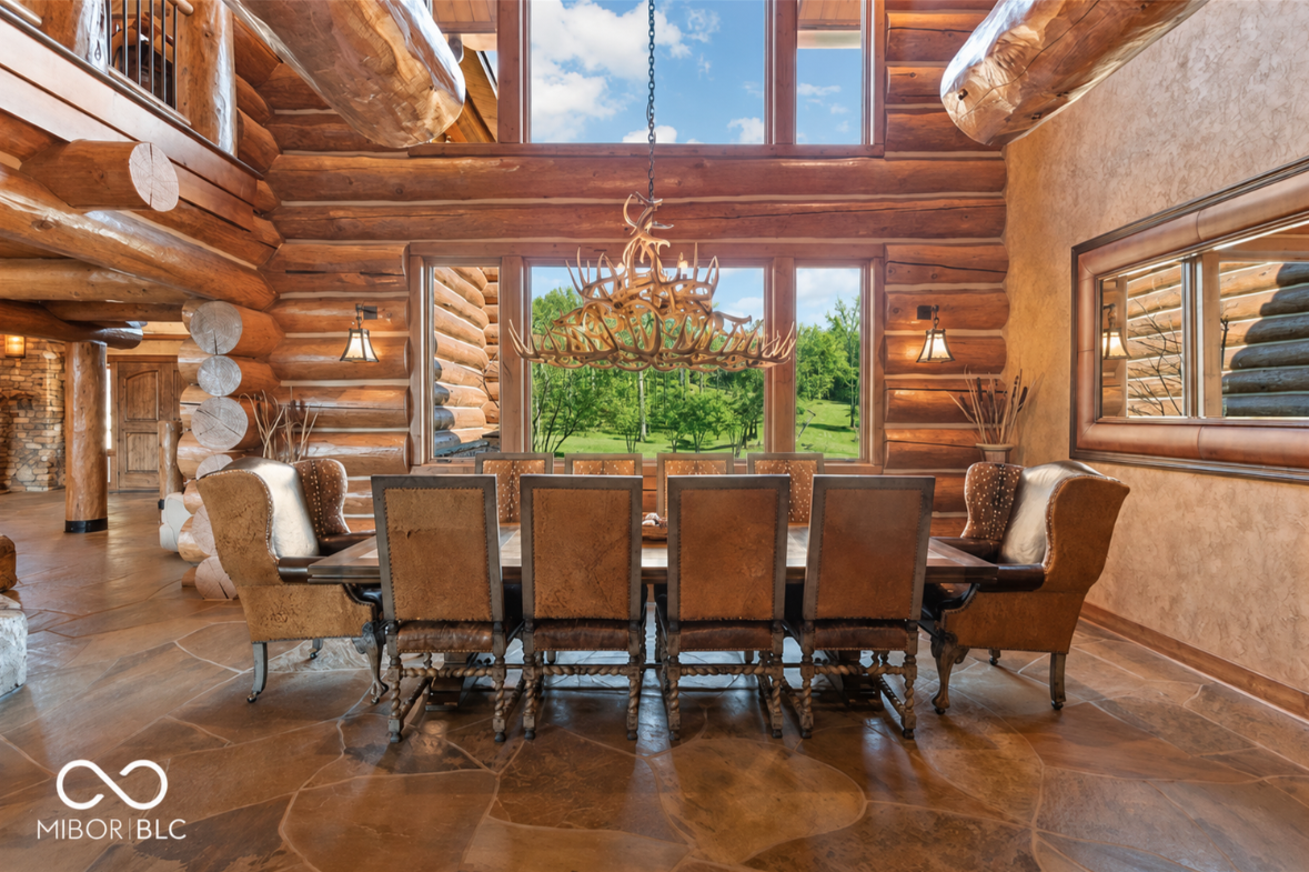 A formal dining room features a long rectangular table that fills the center of the room, surrounded by high-backed leather chairs and positioned directly beneath a large antler chandelier.