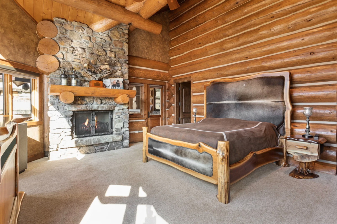 Bedroom with exposed log walls, timber beams, and a stone fireplace across from the bed.
