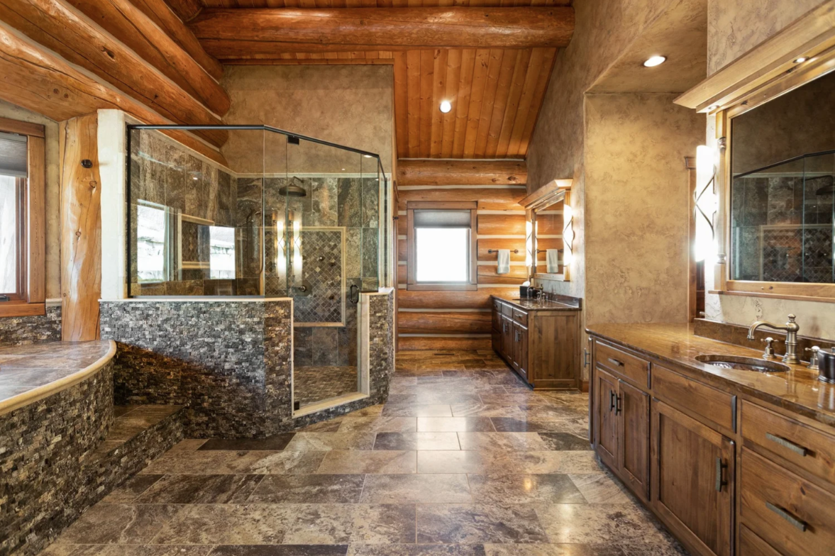 The suite's bathroom opens into a large space finished with stone flooring and wood cabinetry, with dual vanities running along one side and a glass-enclosed shower positioned across the room.