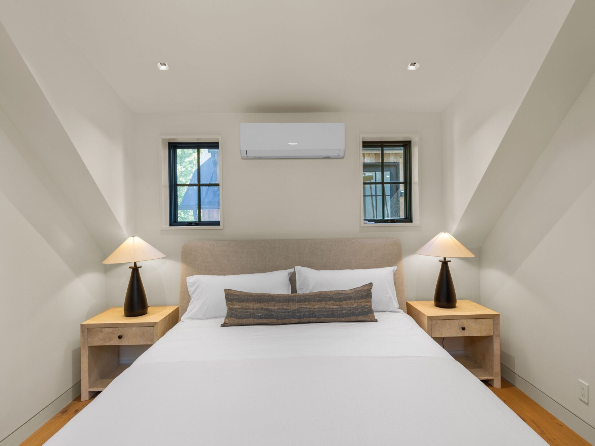 The bedroom sits beneath a sharply pitched ceiling with light walls, wood flooring, and a pair of narrow black-framed windows positioned behind the bed.