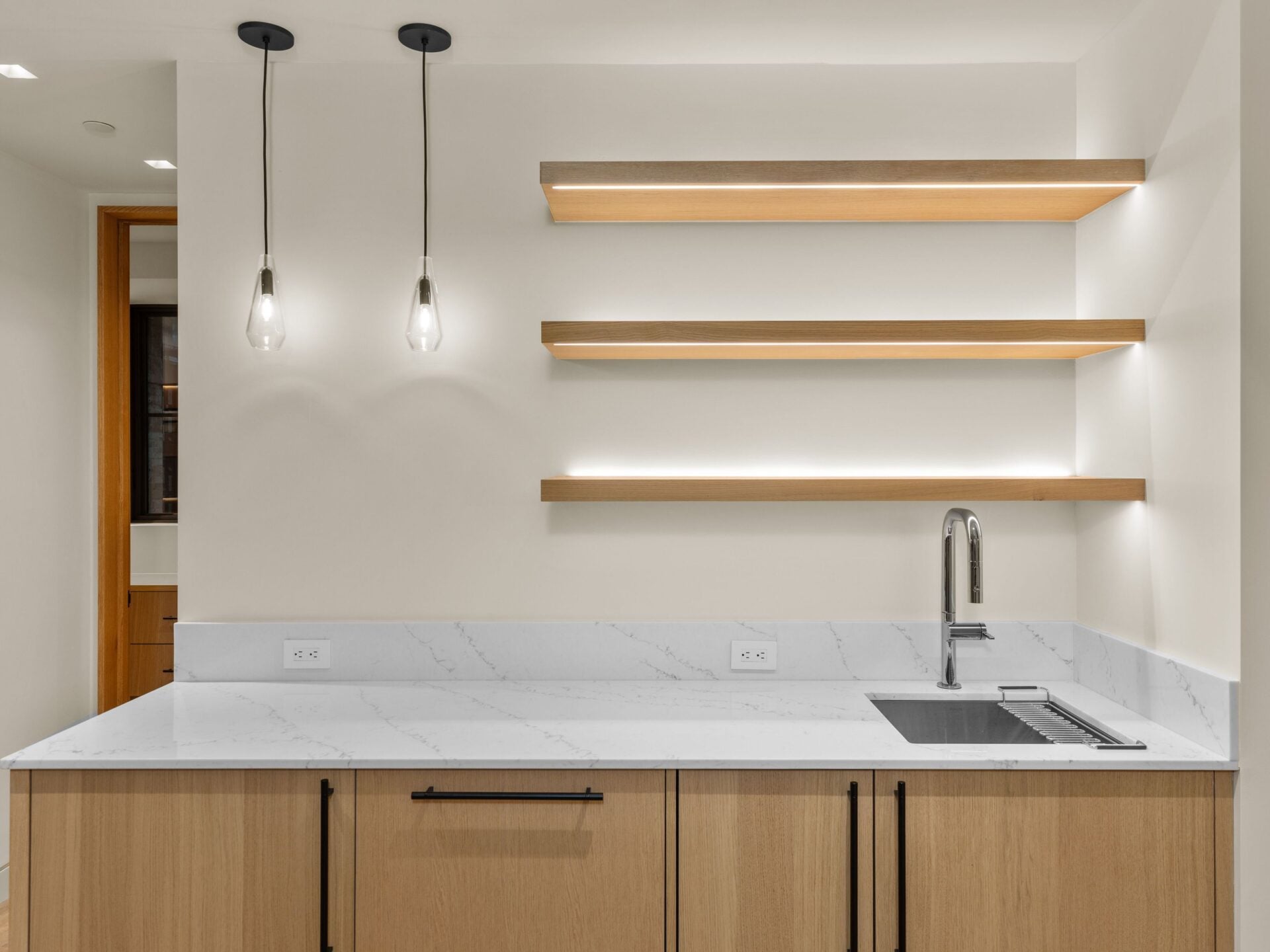 A compact bar offers space to store movie snacks and drinks with stone counters, wood cabinetry, a sink, and floating shelves illuminated from below.
