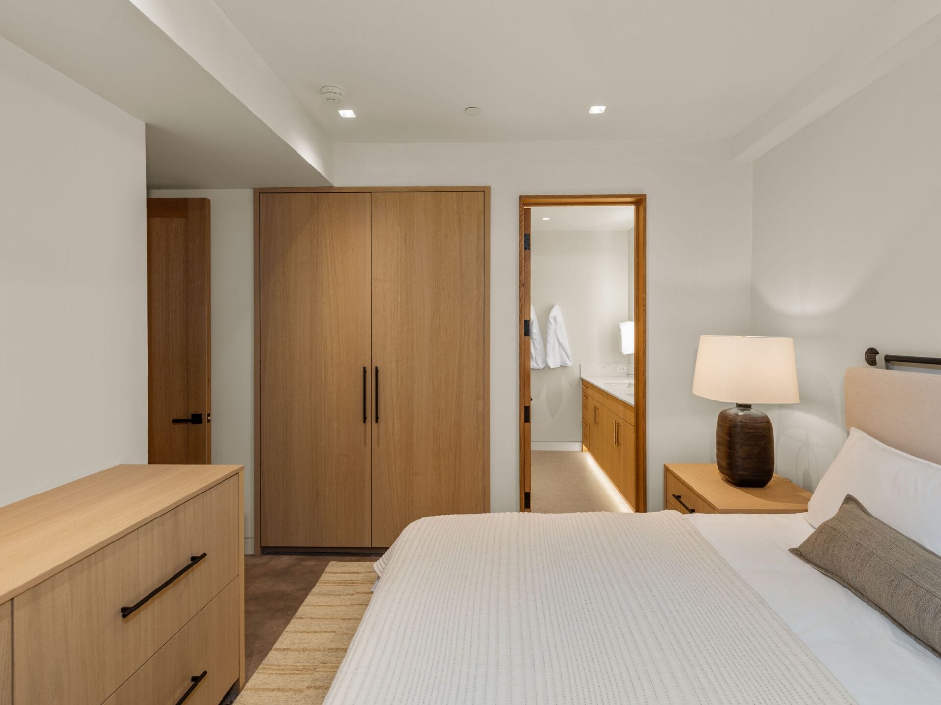 An additional bedroom in the main house continues the same warm yet minimalist design, with light wood cabinetry built directly into the wall and a doorway leading into its private en-suite bathroom.