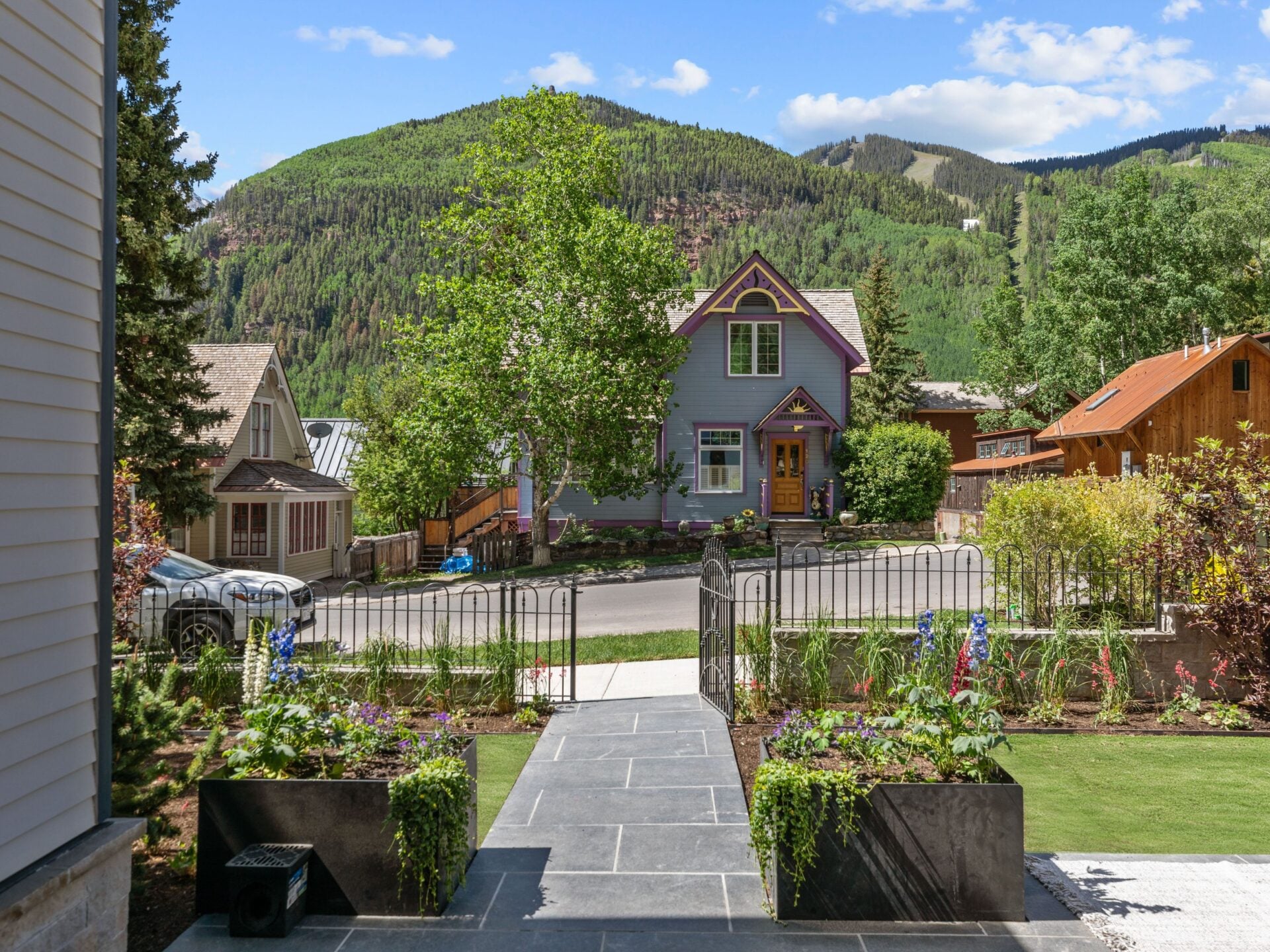A black metal gate opens to a stone walkway that cuts through the landscaped front garden, passing raised planters filled with flowers.