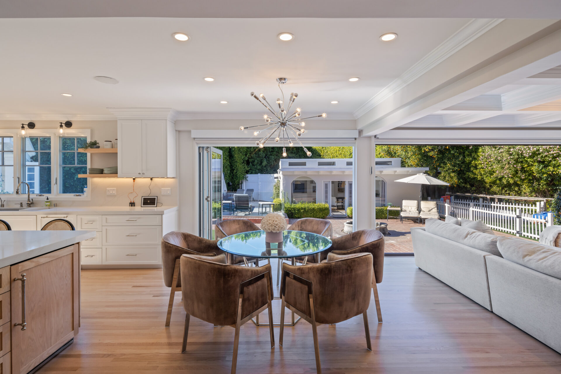 Beside the island, a casual dining area sits beneath a modern Sputnik chandelier, with wide folding glass doors opening along the back wall to the pool and patio.