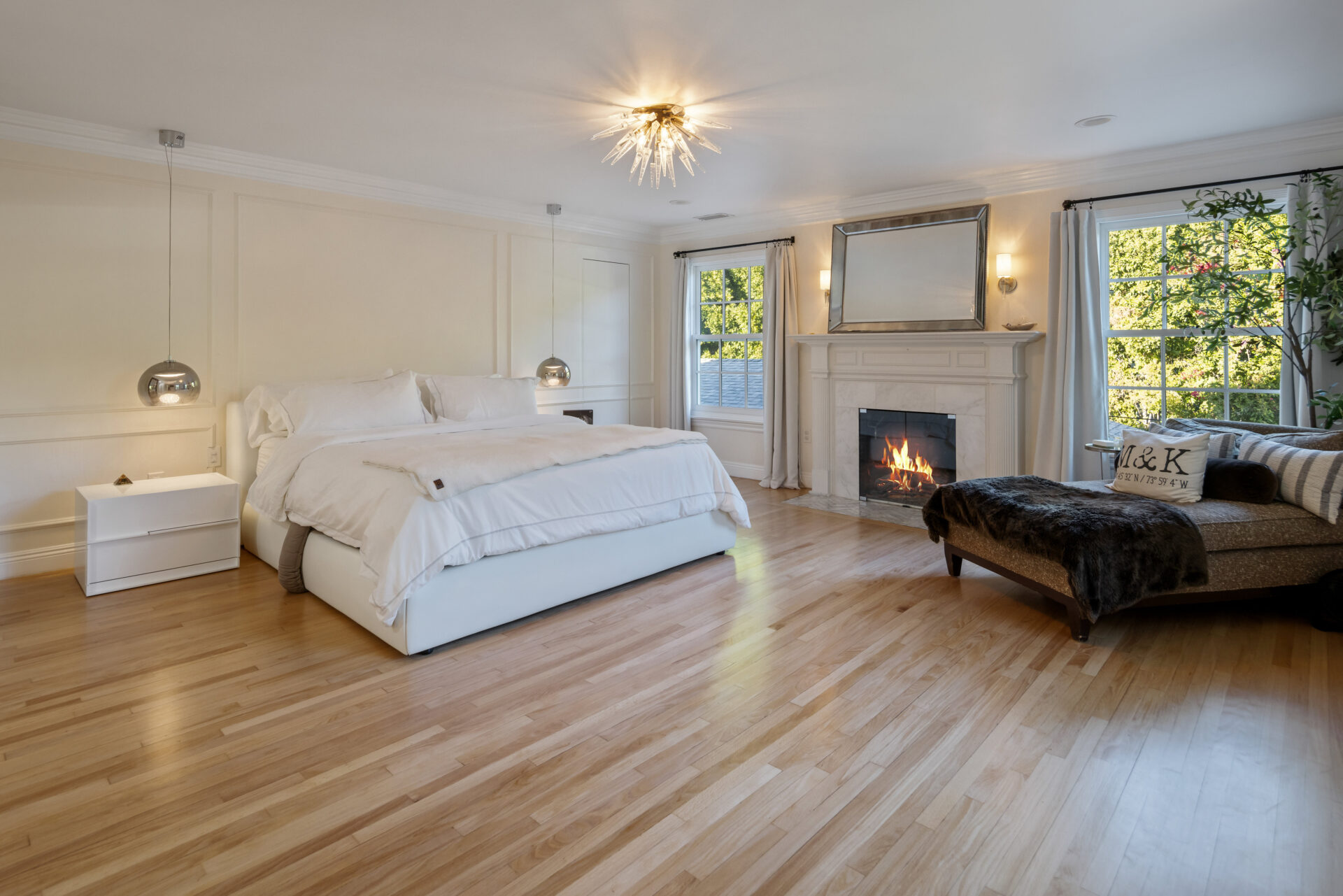 On one side of the bed, a fireplace sits between two large windows, while a chaise creates a separate sitting area alongside it.