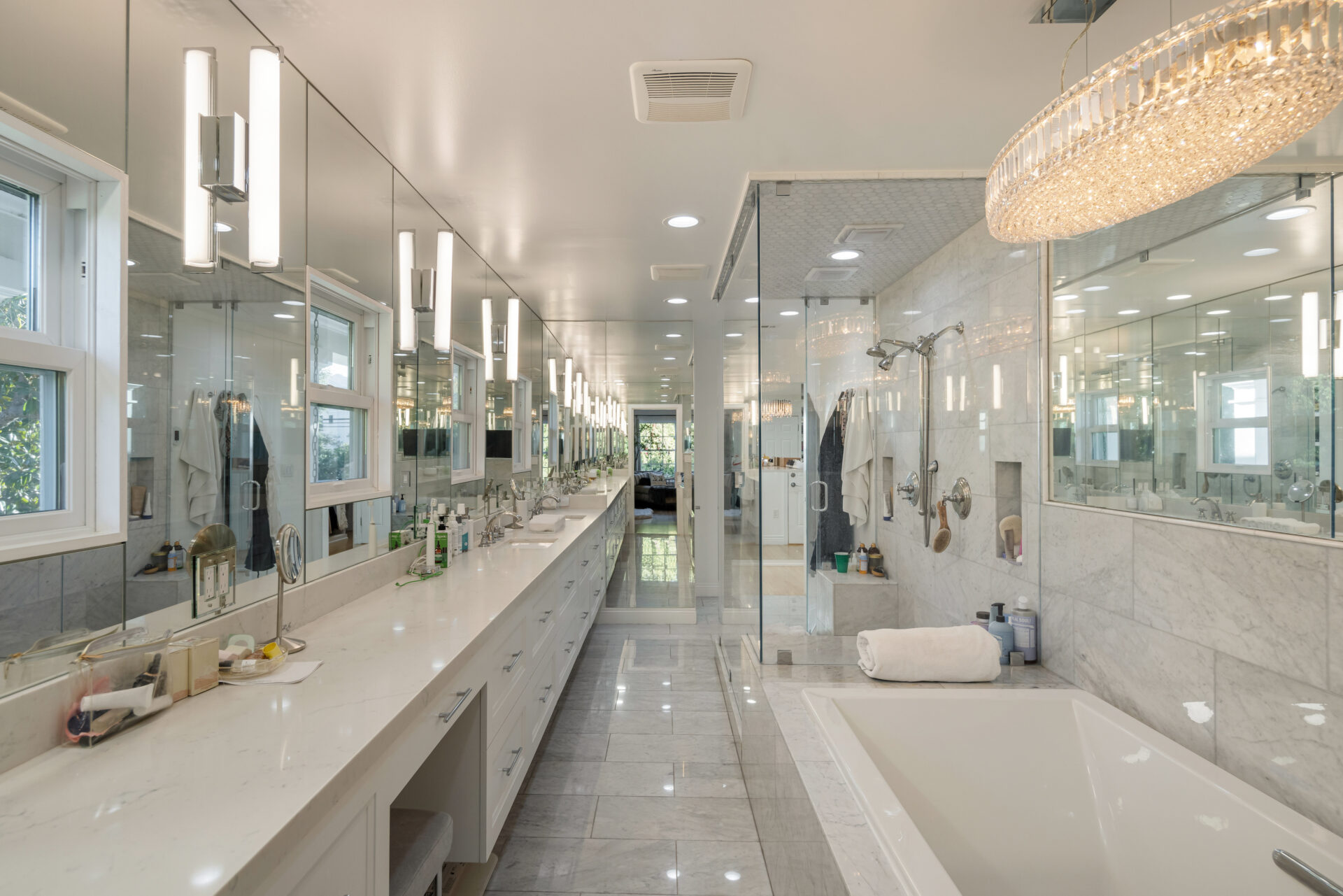 Along one side of the long space, a vanity stretches beneath a continuous mirrored wall, while the opposite side contains a deep soaking tub and a large glass-enclosed shower.