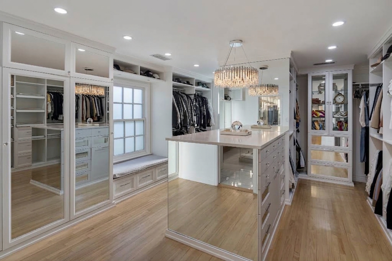 Oversized walk-in closet with built-in storage along both sides with open hanging sections, drawers, upper cabinets, mirrored doors, and glass-front display cabinetry.
